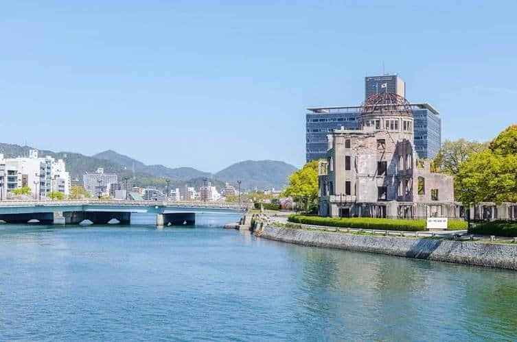 The historical Atomic Bomb Dome ruins standing along the Motoyasu River bank in Hiroshima, Japan, with the Motoyasu bridge and mountains in the background.
