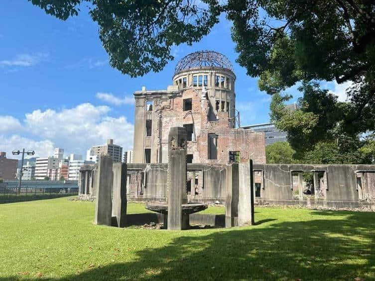 The Atomic Bomb Dome ruins in Hiroshima, Japan, viewed from across the green lawns behind a concrete fountain monument, framed by lush green trees.