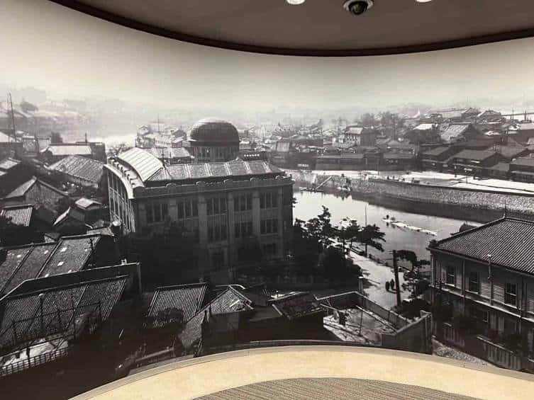 A black and white historical panoramic exhibit inside the Hiroshima Peace Memorial Museum showing the bustling city streets, river, and the intact Prefectural Industrial Promotional Hall before the 1945 atomic bombing.