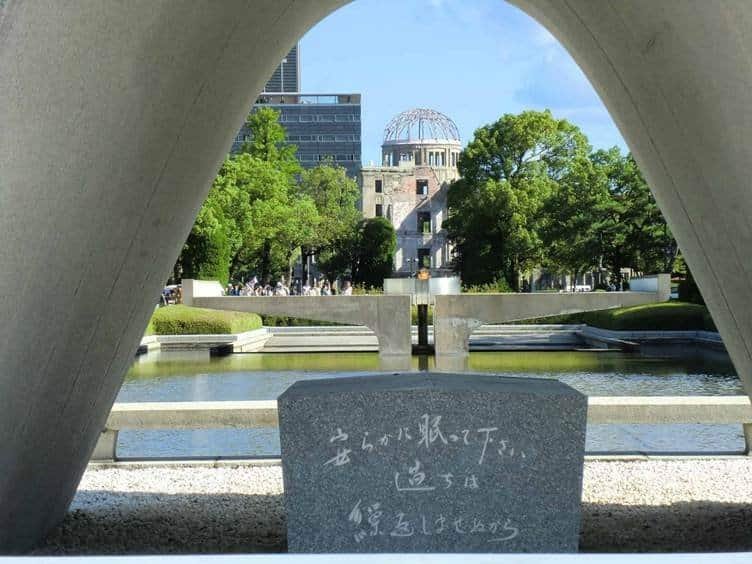 Close-up view framed by the Hiroshima Memorial Cenotaph arch, highlighting the stone monument inscribed with Japanese kanji characters, looking out toward the Peace Flame and the ruined Genbaku Dome under a clear blue sky.