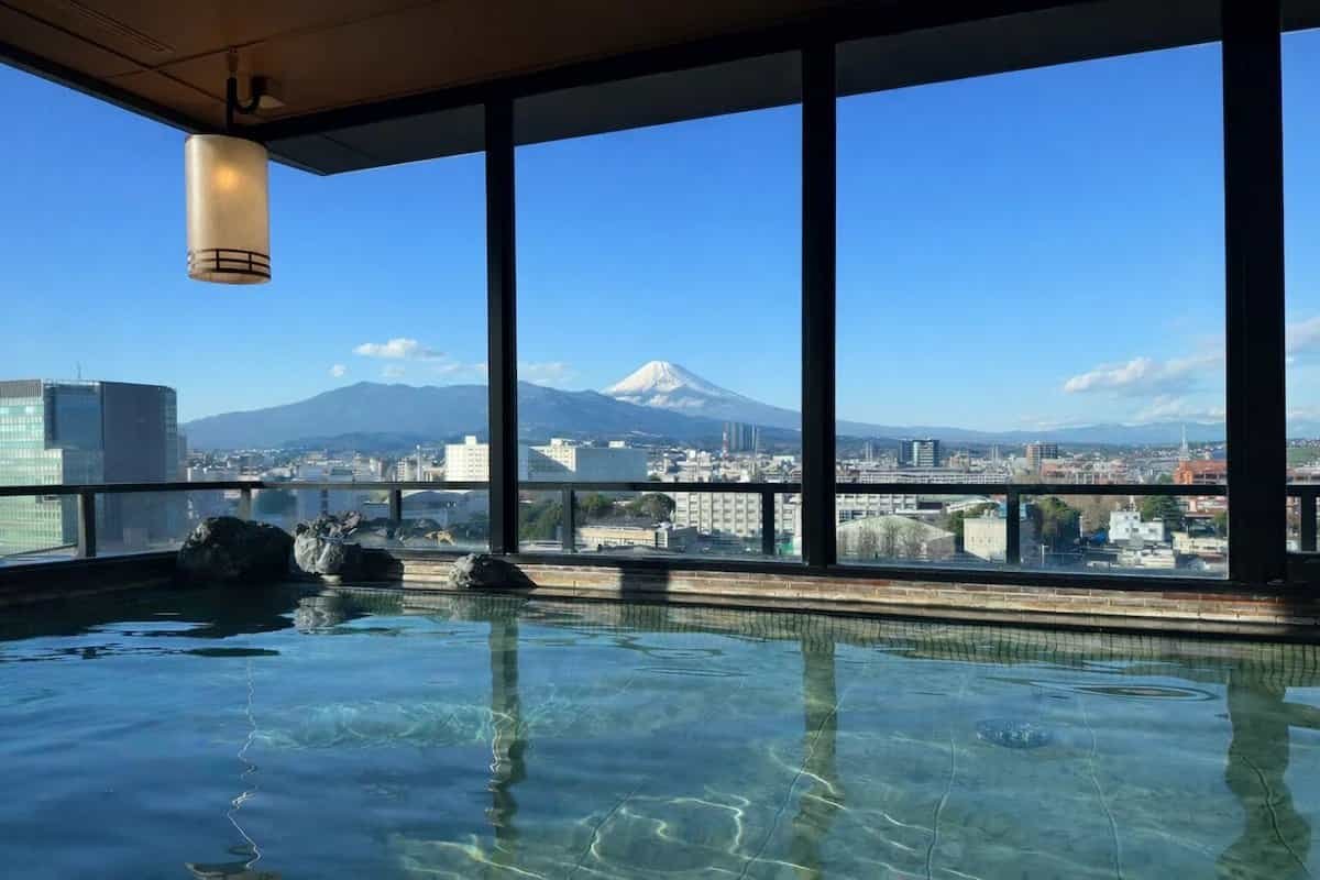 A relaxing indoor public onsen bath at Dormy Inn Mishima in Mishima City featuring large floor-to-ceiling windows with a clear view of Mount Fuji over the city skyline.