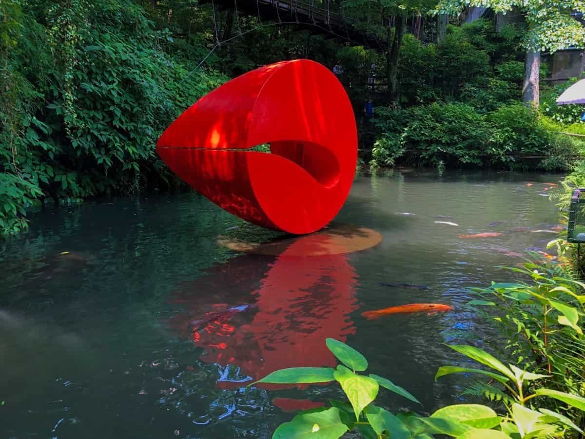 A striking large red abstract sculpture sitting right above a reflective pond with swimming koi fish at the Hakone Open-Air Museum.