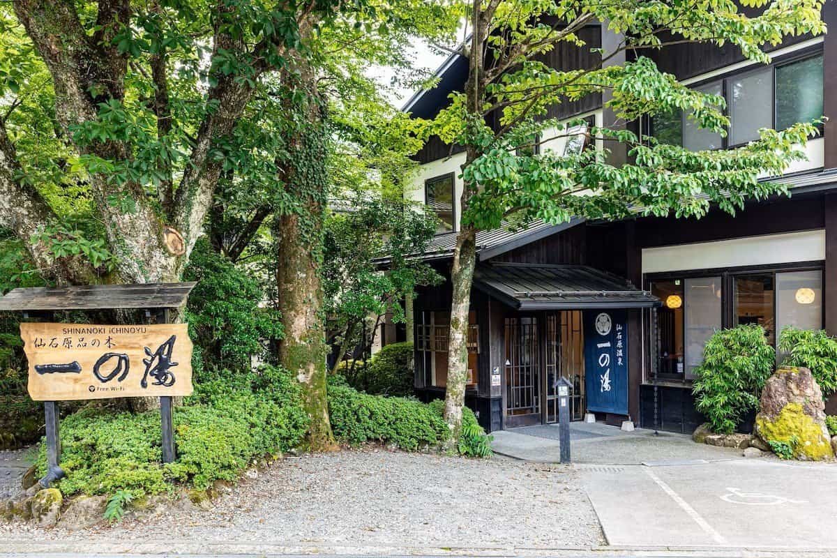 The exterior entrance and wooden sign of Sengokuhara Shinanoki Ichinoyu ryokan in Hakone, surrounded by lush trees and traditional Japanese architecture.