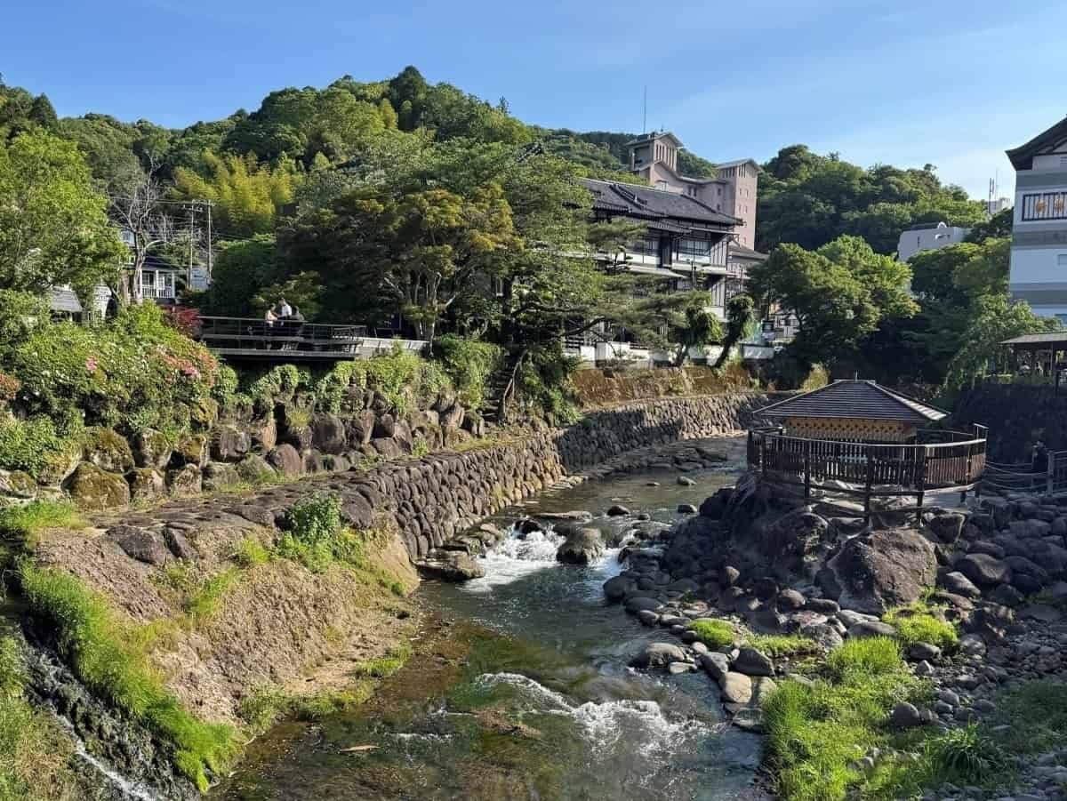 A scenic view of the traditional hot spring town of Shuzenji Onsen, showing historical wooden ryokans, lush green hills, and a river flowing through the center.
